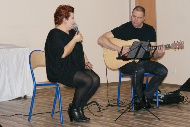 kobieta siedzi na krześle i śpiewa a obok na krześle mężczyzna gra na gitarze