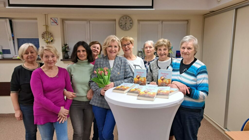 Na zdjęciu widoczne są uczestniczki spotkania ze stojącą po środku autorką książki " Sanatorium uczuć..." panią Moniką Zajączkowską . Osoby stoją wokół stolika z książkami.