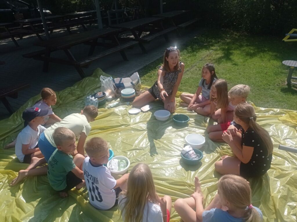 Na zdjęciu widzimy grupę dzieci siedzących w kręgu na rozłożonej plastikowej płachcie na trawie, podczas słonecznego dnia. Dzieci biorą udział w zajęciach sensorycznych, podczas których będą robiły piasek kinetyczny z użyciem skrobi kukurydzianej i oleju. Instruktorka siedzi pośrodku, wskazując na materiały, a dzieci z uwagą obserwują i przygotowują się do działania.