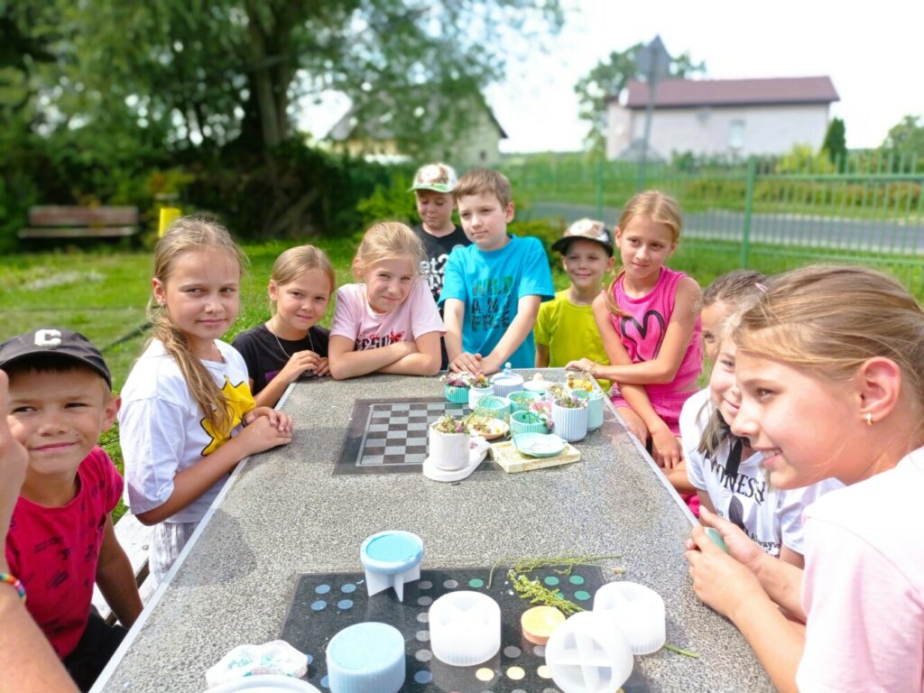 a zdjęciu widać grupę dzieci siedzących przy stole na świeżym powietrzu.