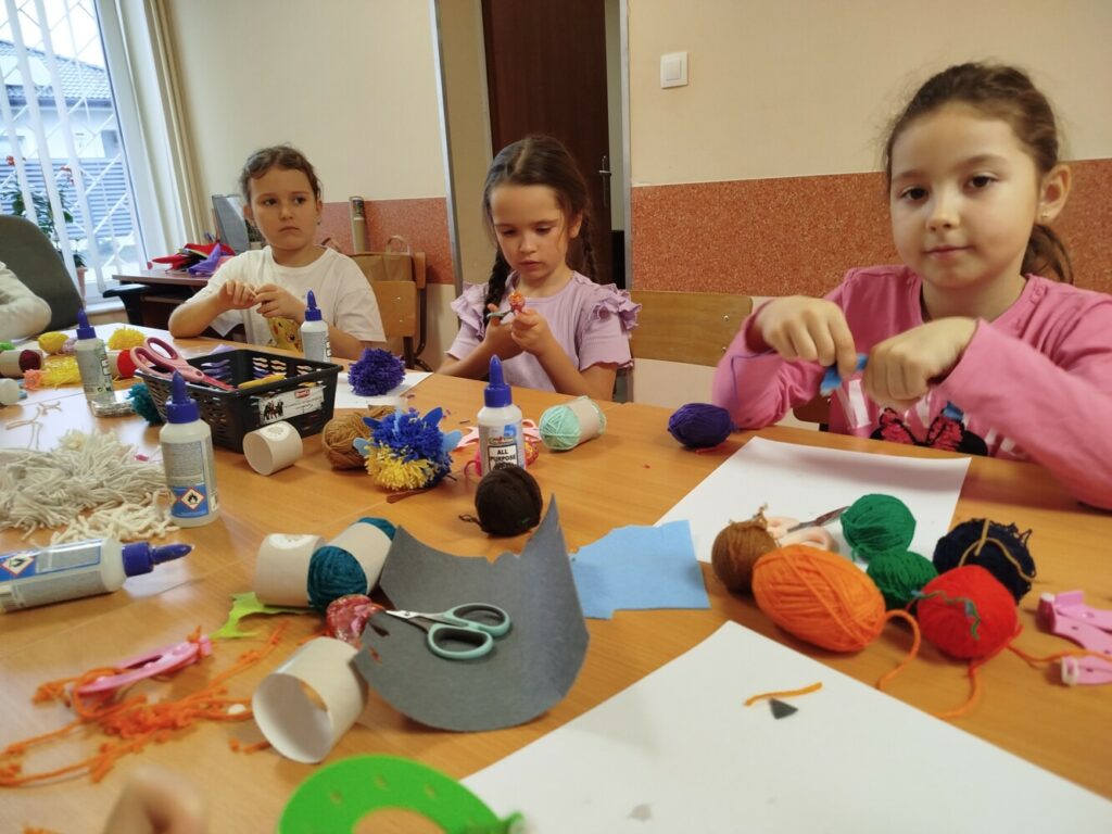 Na zdjęciu dzieci siedzą przy stole i tworzą pompony z kolorowej włóczki. Przed nimi leżą kłębki włóczki, kleje, nożyczki i inne materiały plastyczne. Dziewczynki skupione są na swojej pracy. Atmosfera jest twórcza i radosna.