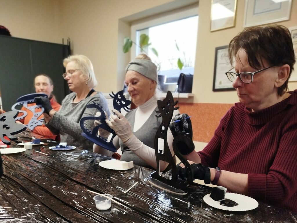 Grupa czterech osób bierze udział w warsztatach rękodzielniczych. Każda z nich maluje drewniane stojaki na biżuterię w kształcie kotów. Stoły przykryte są czarną folią ochronną, a na talerzykach widać farby i pędzle. Uczestniczki są skupione na swojej pracy, a atmosfera wygląda na pełną zaangażowania i kreatywności.